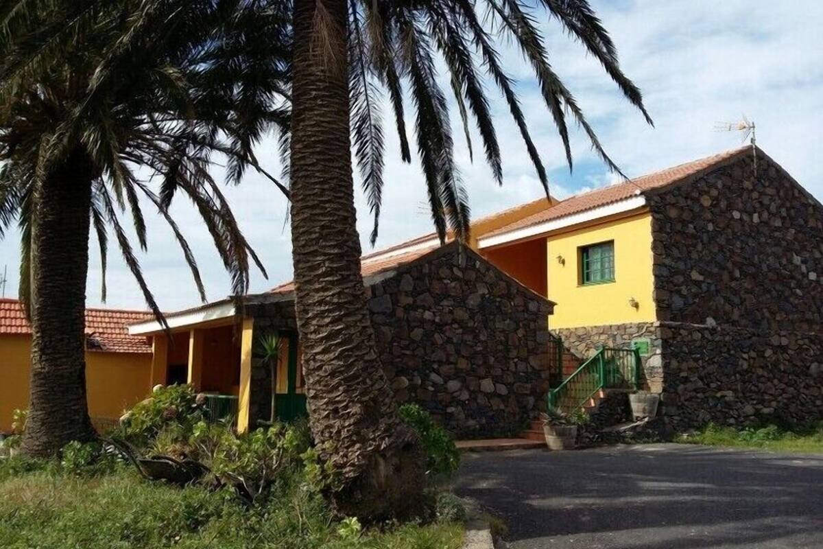 A two-story building with a stone facade and a yellow upper section. There is a staircase leading to the upper floor, and a small garden area with palm trees and shrubs.