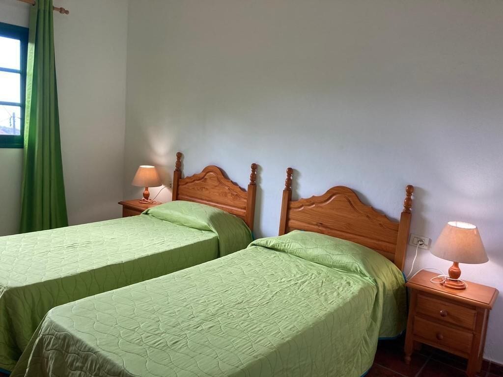 Habitació amb dos llits individuals, amburts de fusta, taules de nit amb làmpades i una finestra amb cortines verdes.