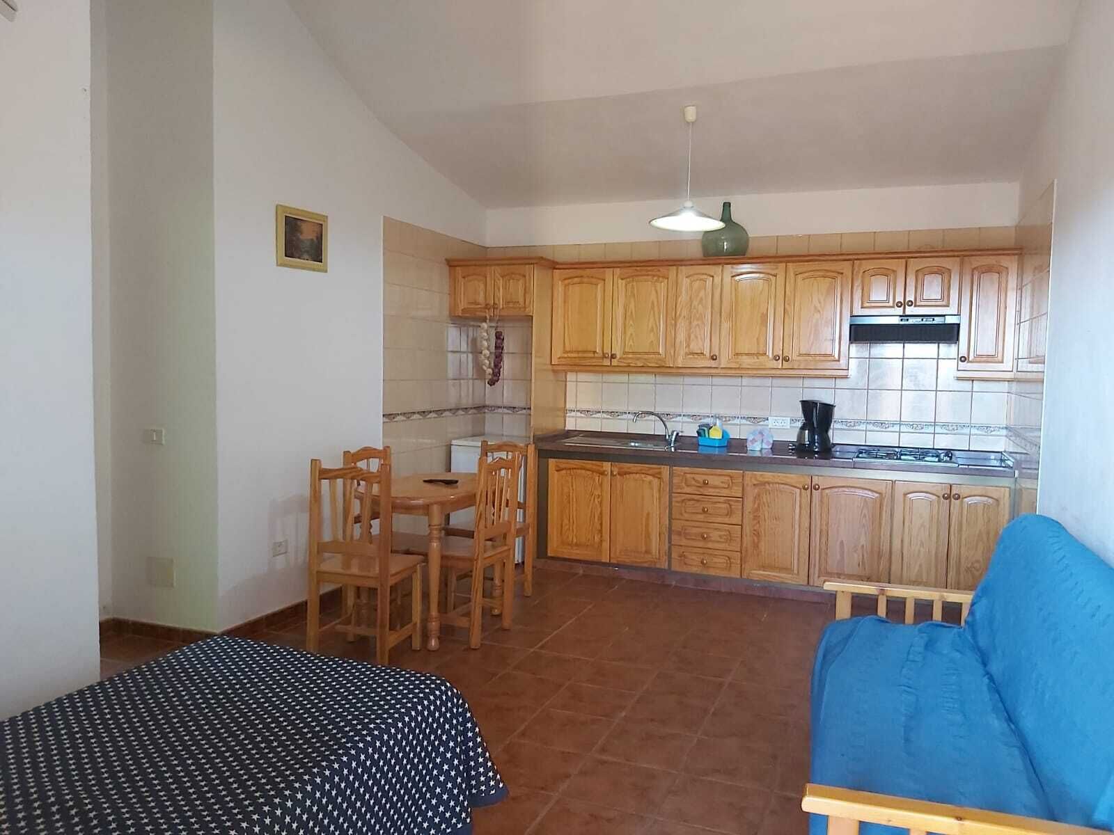 A compact kitchen with wooden cabinets, a dining table with chairs, a blue sofa, and a hanging light fixture.