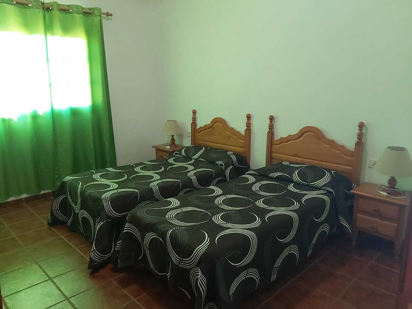 A room with two beds, green curtains, wooden headboards, bedside tables with lamps, and a tiled floor.