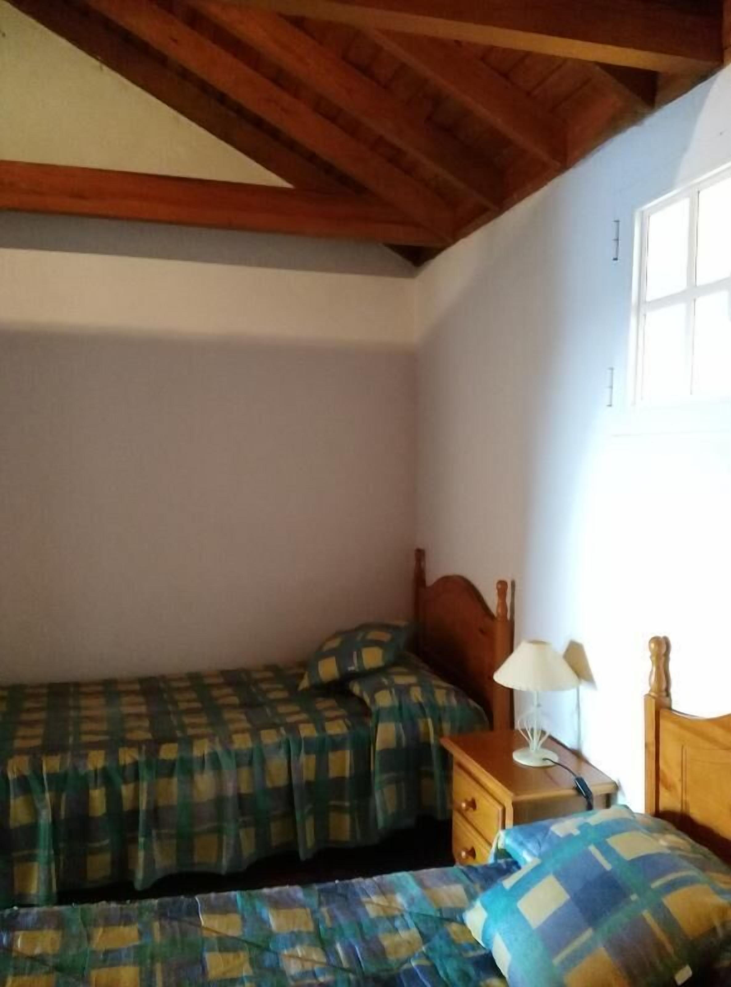 A bedroom with two beds, wooden headboards, a bedside table with a lamp, and a window with curtains.