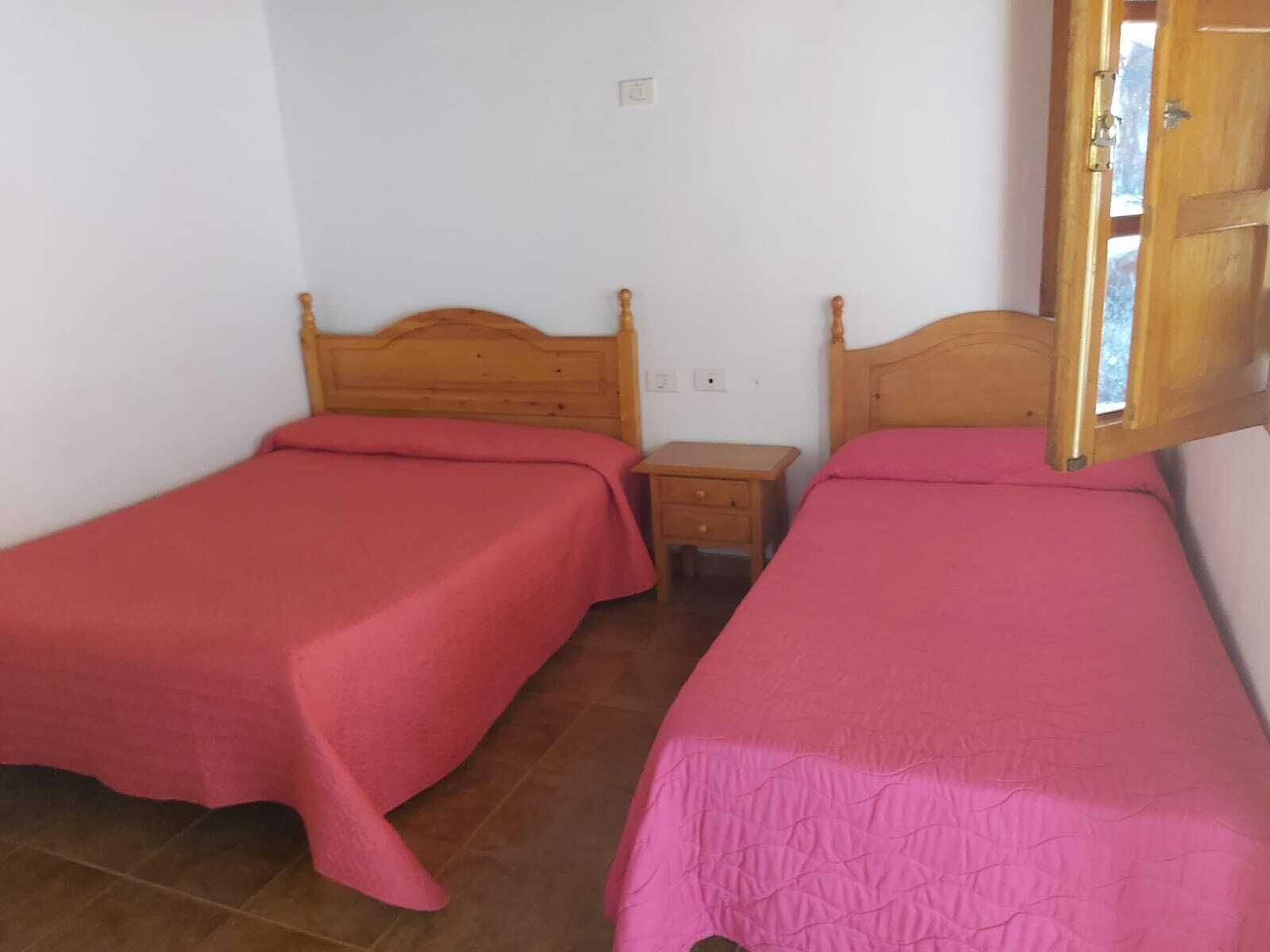 Une petite chambre avec deux lits simples, chacun doté d’une tête de lit en bois et d’une couvre-lit rose. Une petite table de chevet en bois se trouve entre les deux lits.