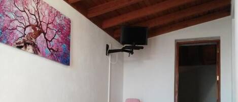A room with a wooden bed, a painting of a tree, a television mounted on the wall, and a lamp on a small table.