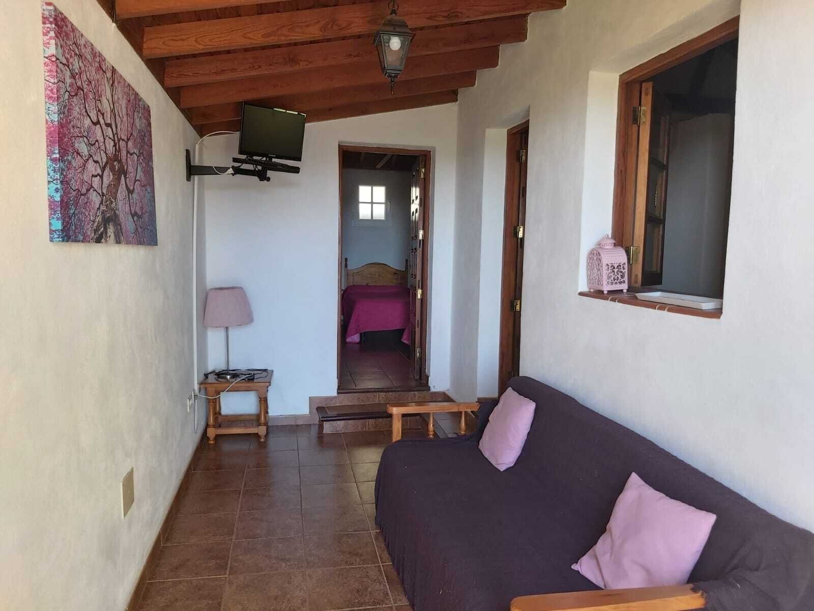 A cozy room with a wooden ceiling, a sofa, a small table with a lamp, and a bed in the background.