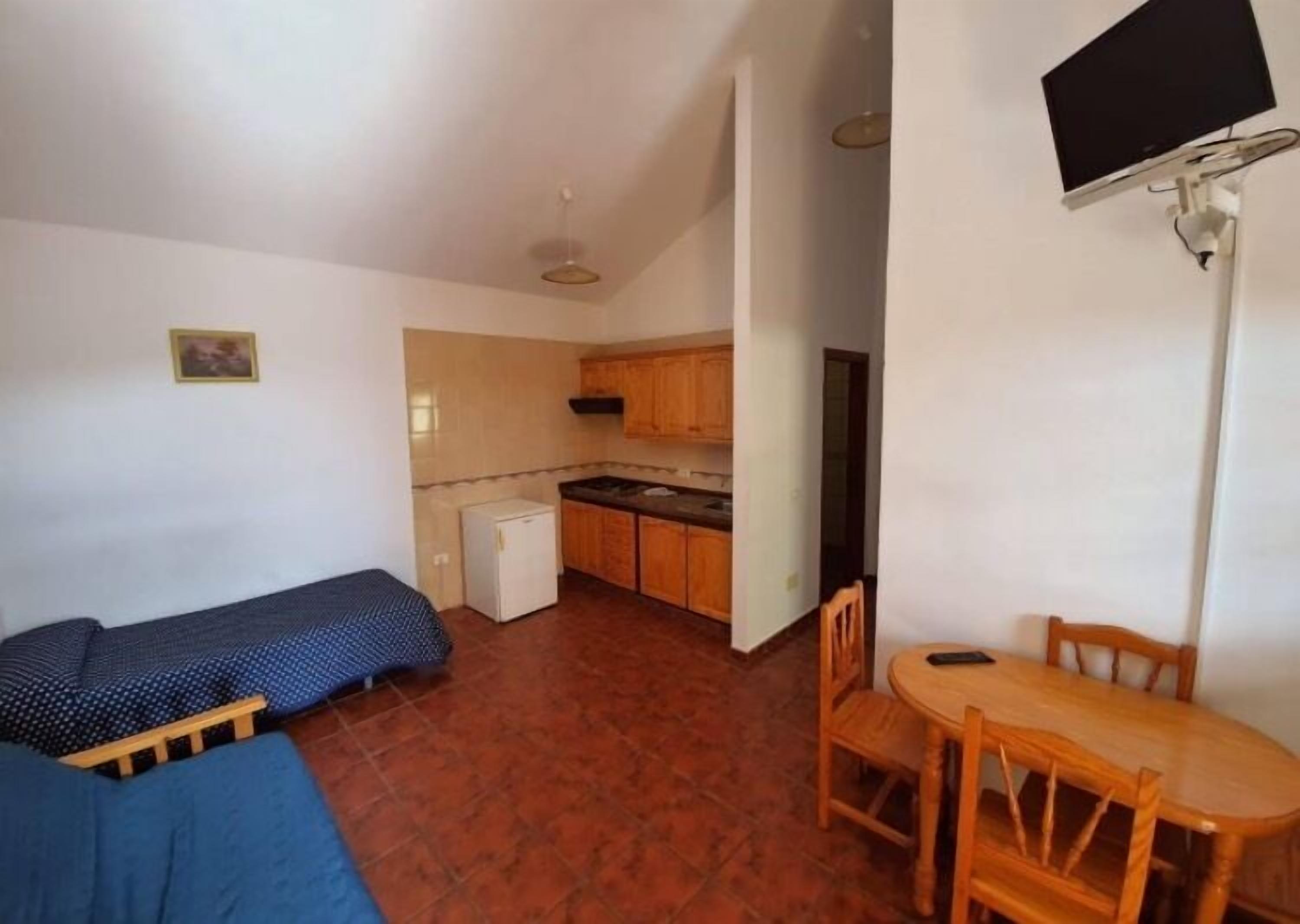 A compact room with a bed, a small kitchenette, a dining table with chairs, and a TV mounted on the wall.