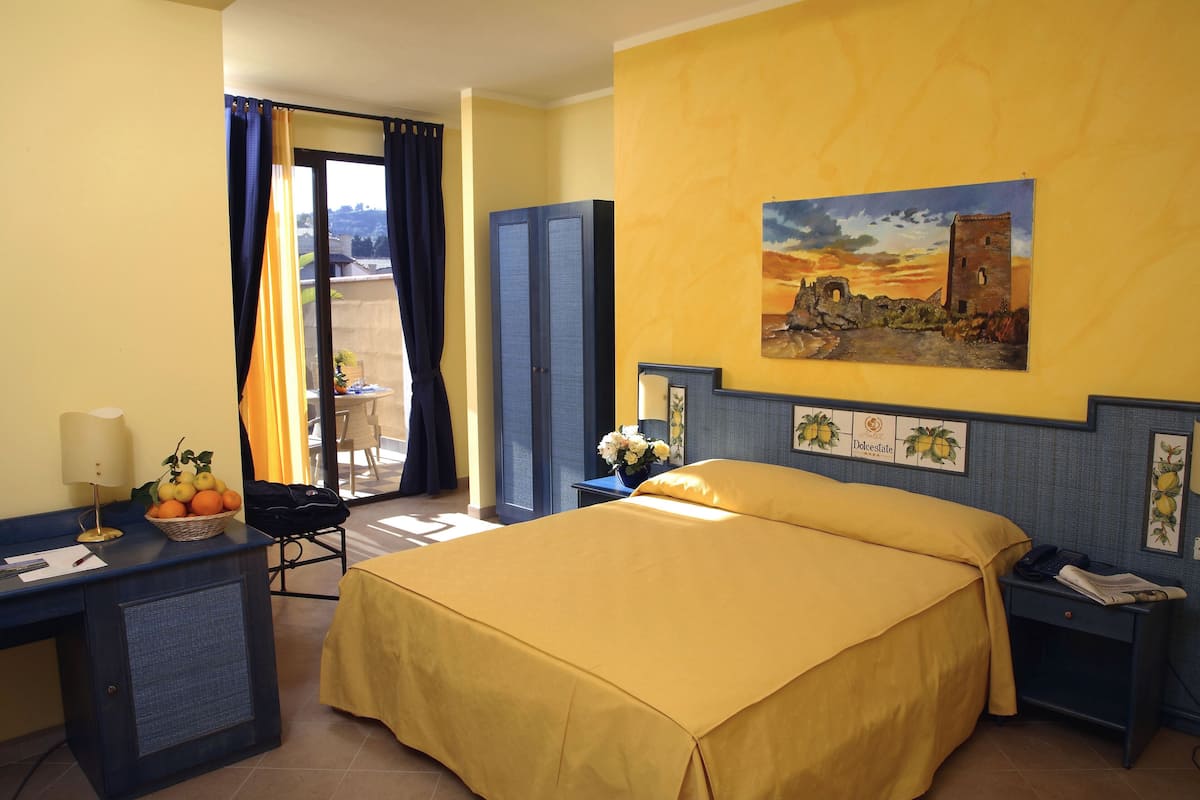Habitació d'hotel amb un llit, una taula de treball amb una làmpada, una cadira, una finestra amb cortines i un quadre a la paret.