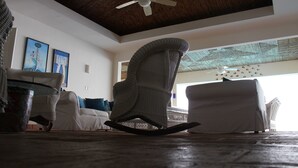 A room with a ceiling fan, a rocking chair, a sofa, and a large window with a view of the beach. - Casa Mallen - La Boquita (Diriamba)