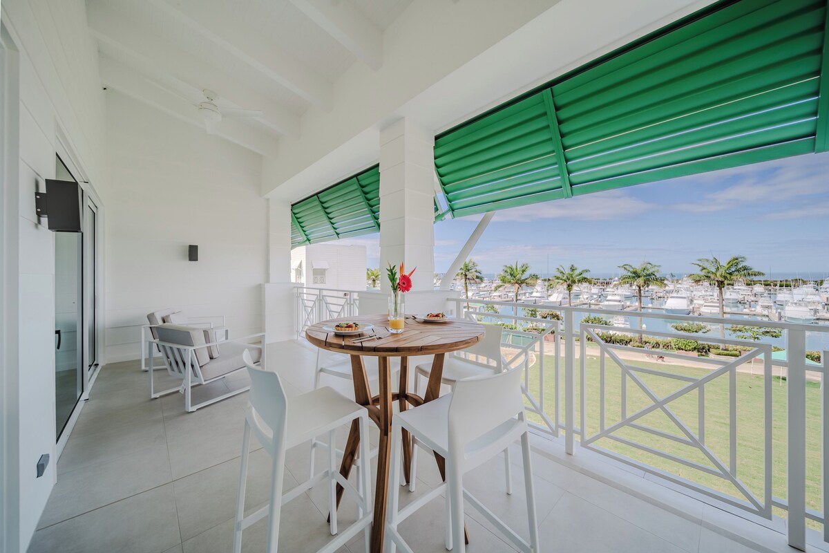 Ein Balkon mit weißer Möbel, einem runden Holztisch und Blick auf eine Marina mit Booten.