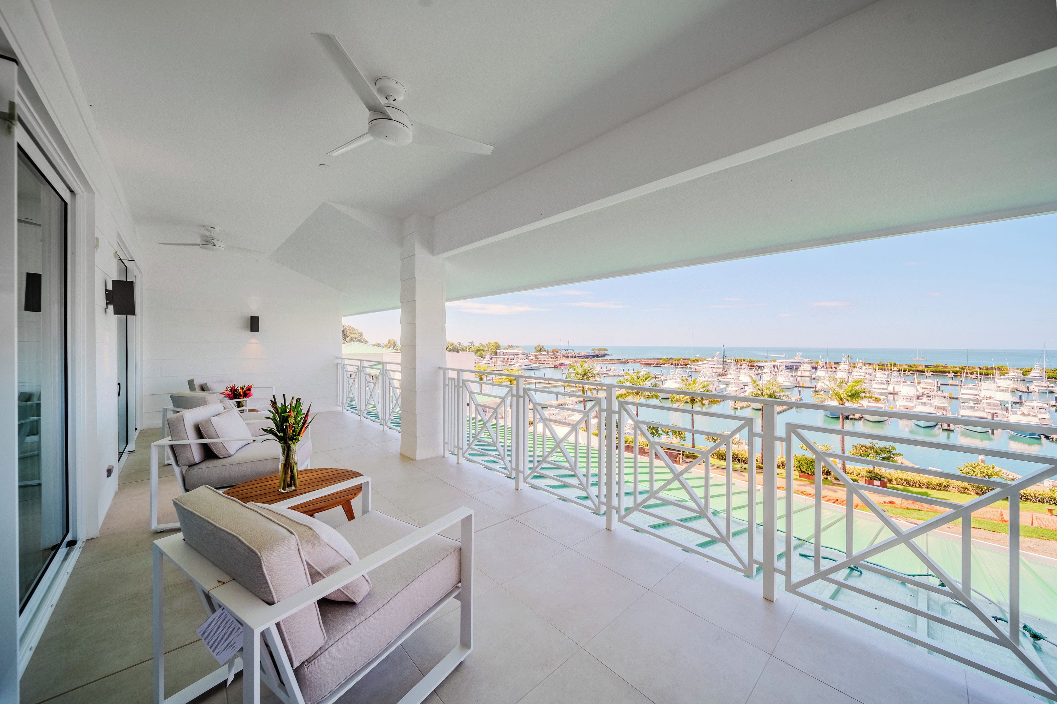 A balcony with a view of the sea and marina, white furniture, and a ceiling fan.