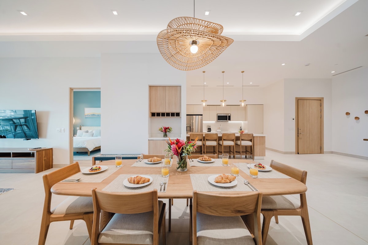 Un comedor moderno con una mesa de madera lista para el desayuno, una cocina al fondo y una sala de estar con un sofá y un televisor.