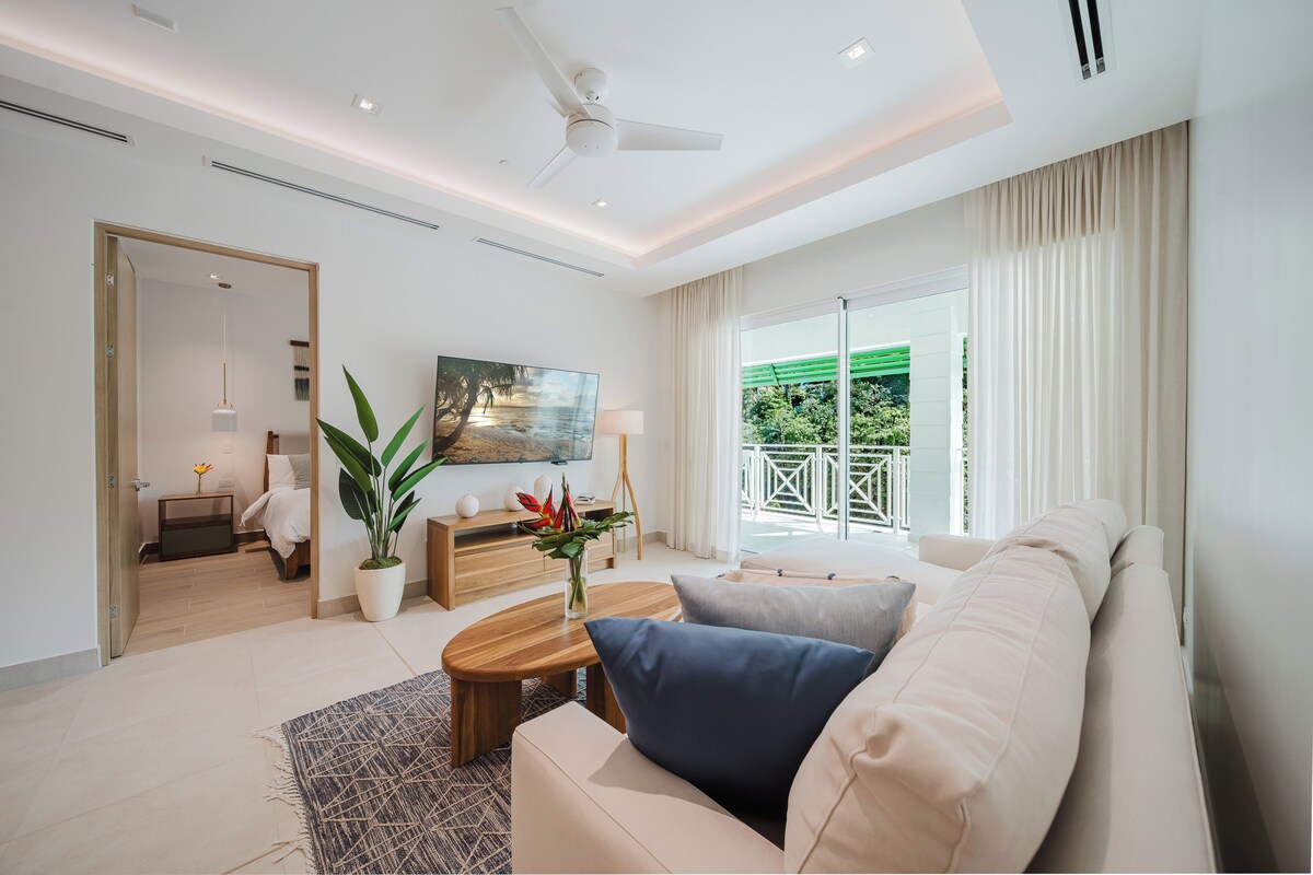 Ein modernes Wohnzimmer mit einer Couch, einem Couchtisch und einem Fernseher. Ein Balkon bietet Blick auf Grünflächen.