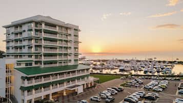 A multi-story hotel with balconies, a parking lot, and a marina with boats in the background.