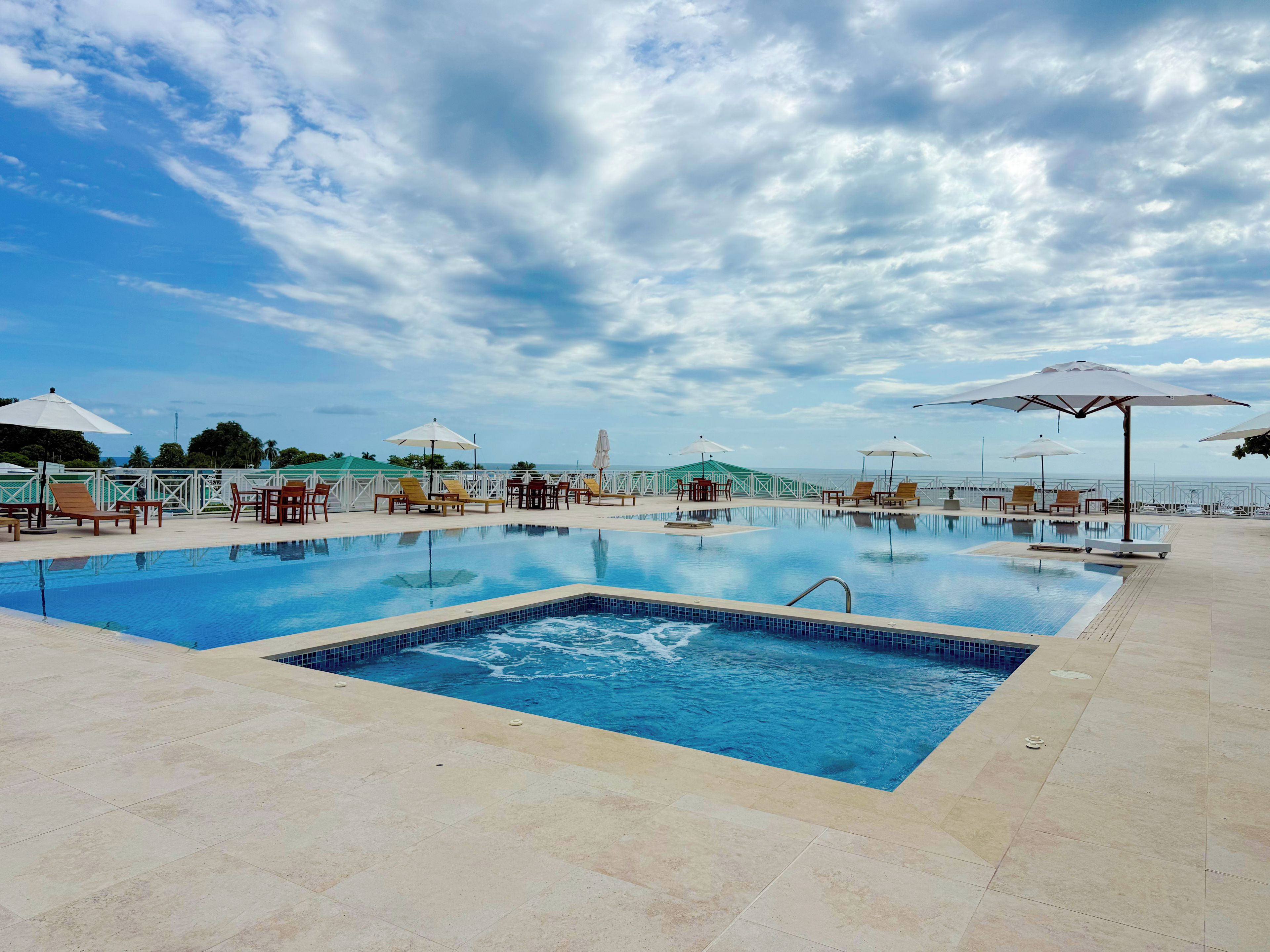 A swimming pool with lounge chairs and umbrellas, surrounded by a fence, under a partly cloudy sky.