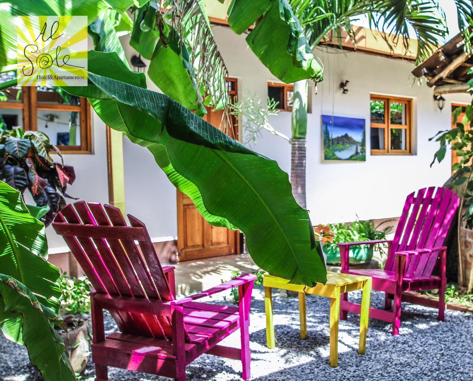 Un patio con dos sillas de madera y una pequeña mesa, rodeado de abundante vegetación y un edificio blanco con ventanas.