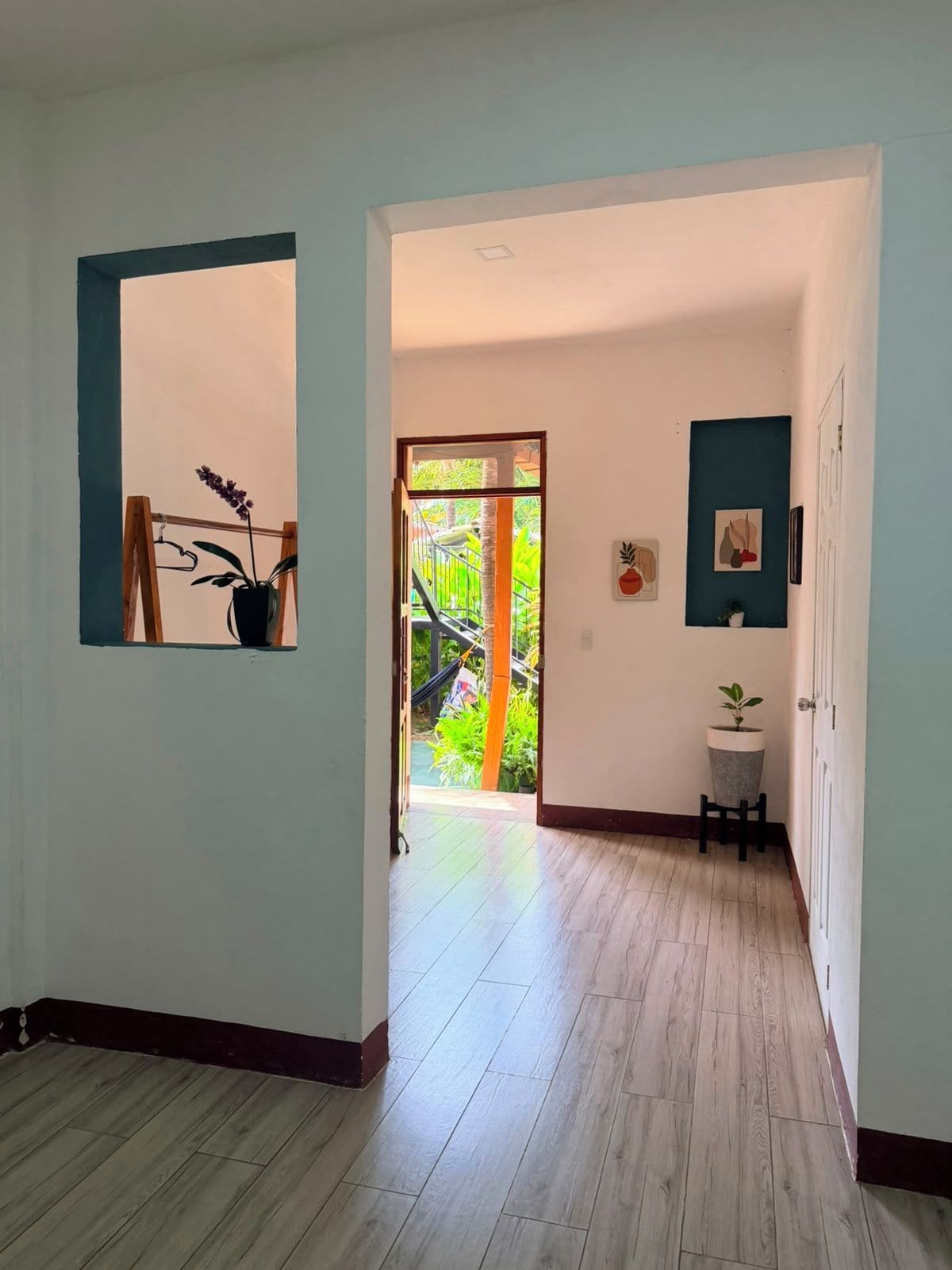 Un pasillo con suelo de madera, una puerta que da a un balcón y una pequeña mesa con una planta.