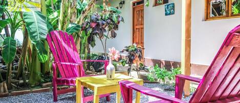 Un patio con sillas y mesa de madera, rodeado de plantas tropicales y un edificio con puerta y ventanas de madera.