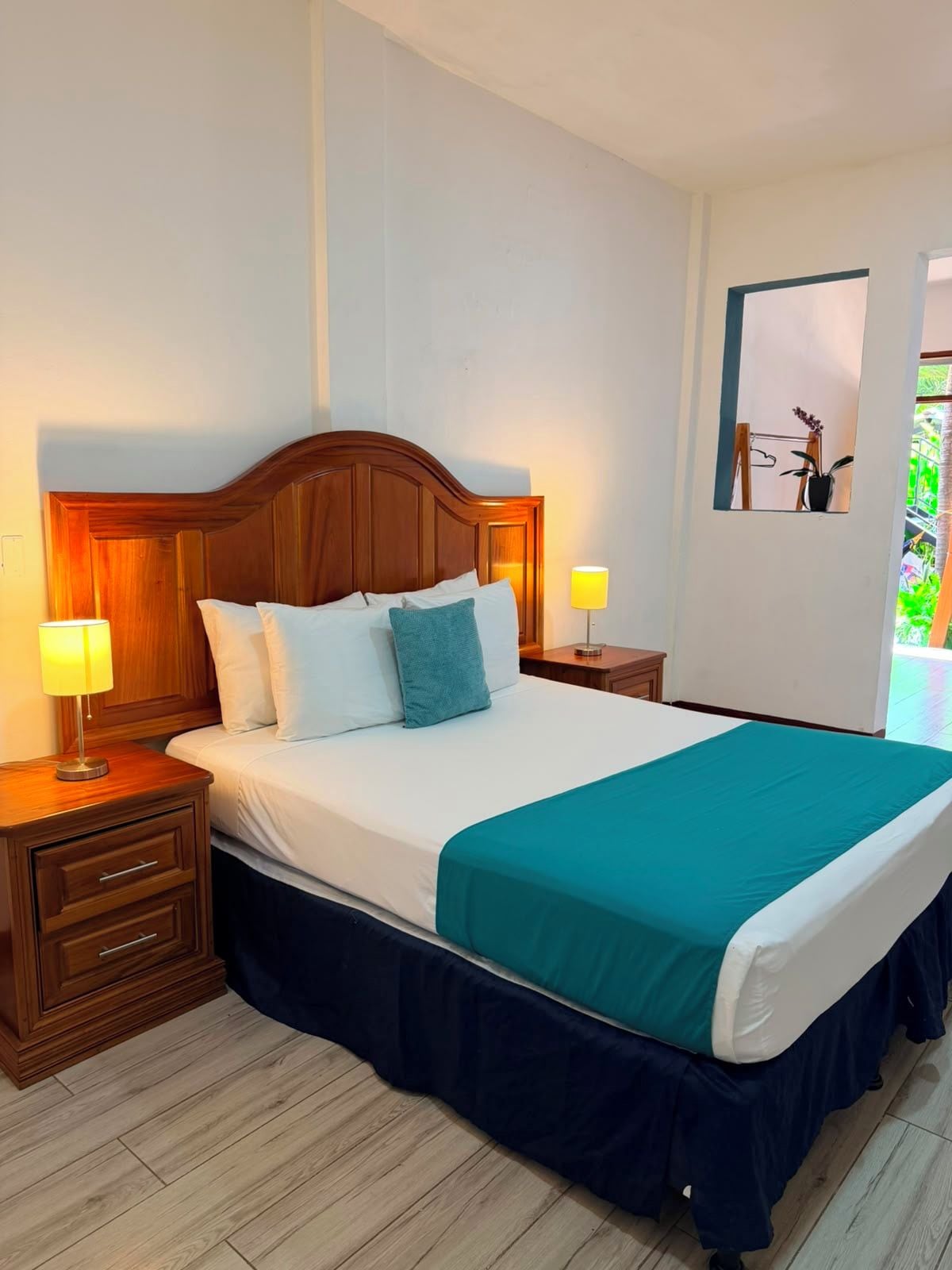 Habitación de hotel con cabecera de madera, cama grande con ropa de cama blanca y aguamarina, dos lámparas de noche, una mesita y una ventana con vistas a la vegetación.
