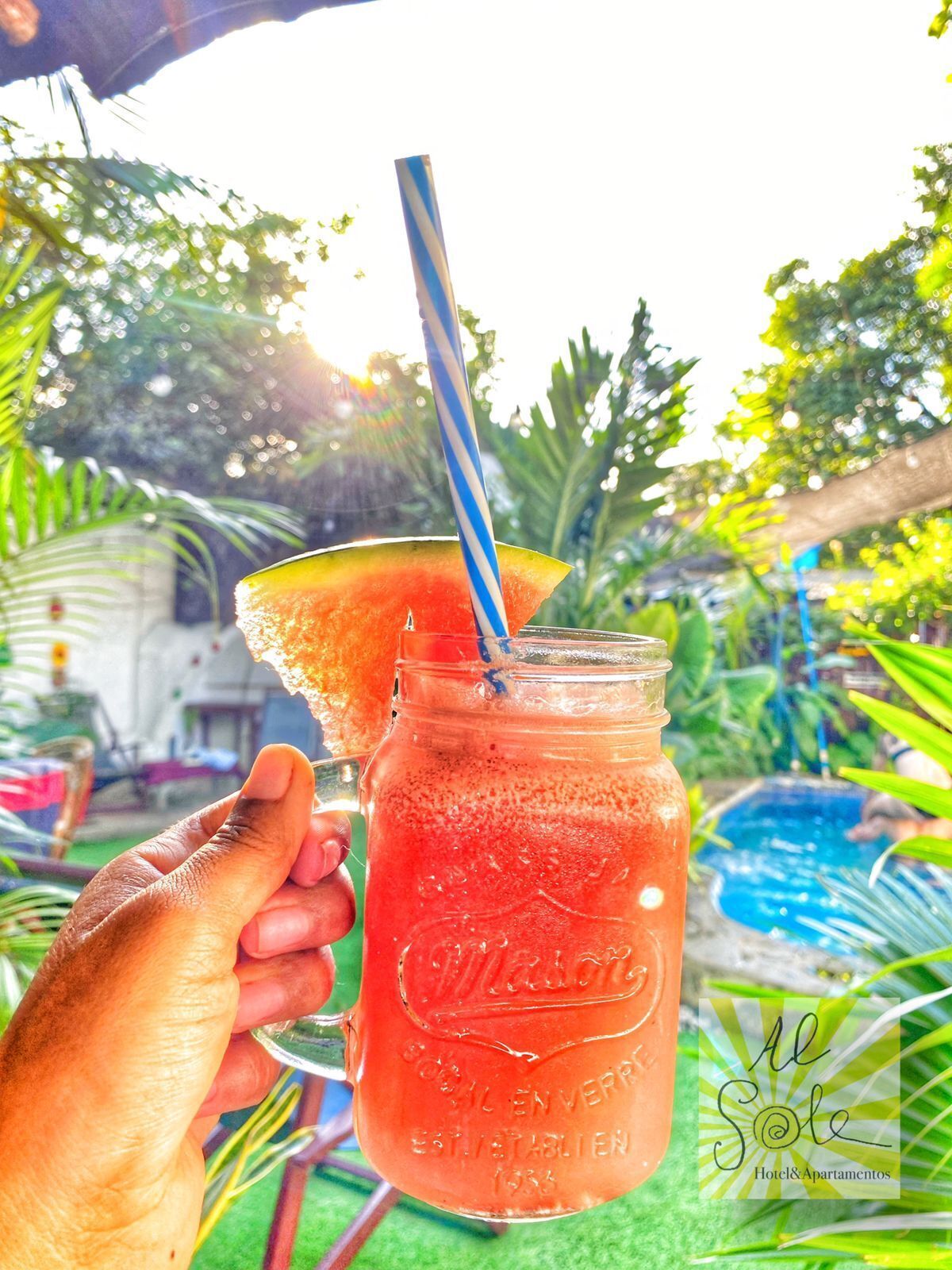 A hand holding a mason jar with a red drink and a slice of watermelon on the rim.