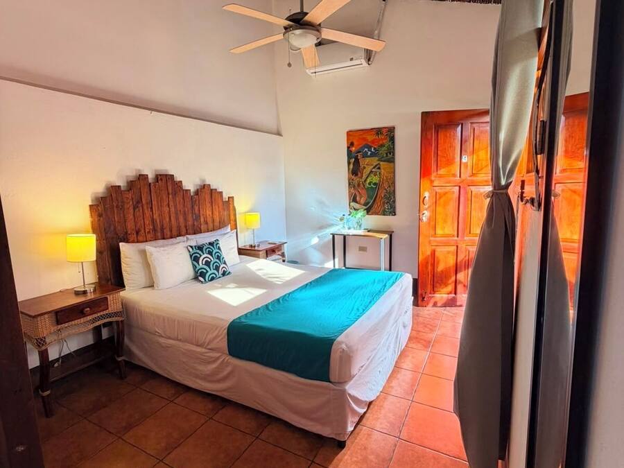 Un dormitorio con cama, mesita de noche, escritorio, ventilador de techo y un cuadro en la pared.