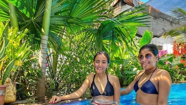 Two people in a swimming pool with tiled flooring, surrounded by lush greenery and a stone building in the background. - Al Sole Guesthouse (León)