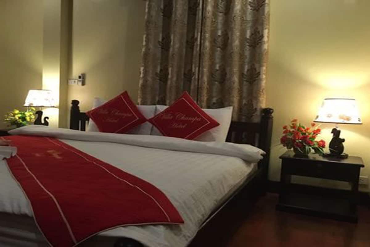 Una cama bien hecha con una colcha y almohadas rojas, una mesita de noche con una lámpara, y un florero con flores.