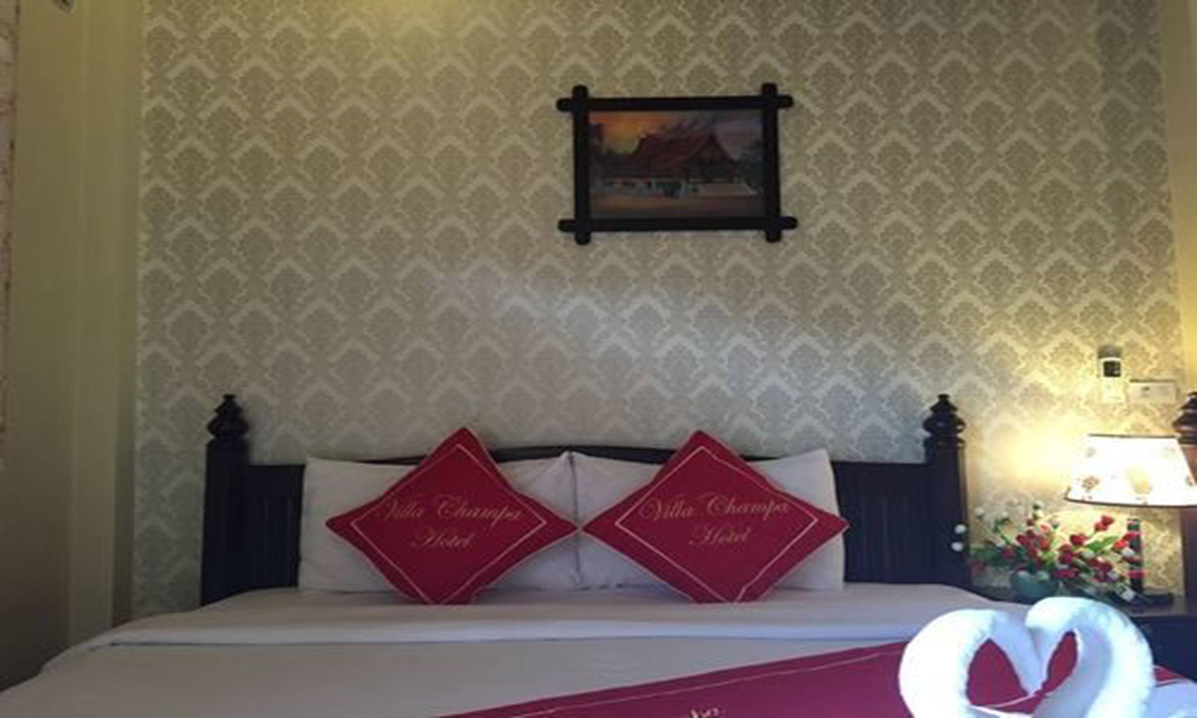 Una habitación de hotel con papel tapiz estampado, un cuadro en la pared y dos almohadas rojas con texto.