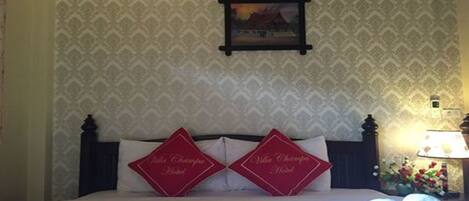 Una habitación de hotel con papel tapiz estampado, un cuadro en la pared y dos almohadas rojas con texto.