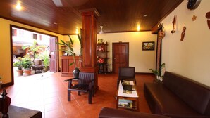 A lobby with a wooden ceiling, a reception desk, a sofa, and several chairs. There are potted plants and a clock on the wall. - Villa Champa (Luang Prabang)