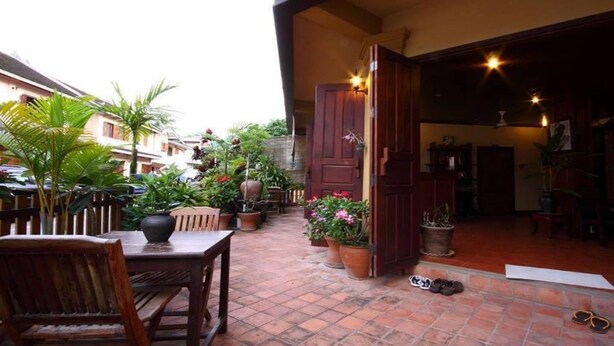 An outdoor patio area with a wooden table and chairs, potted plants, and a red door leading to an interior space. - Villa Champa (Luang Prabang)