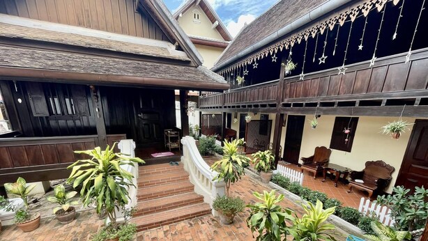 Un patio con piso de ladrillo, plantas en macetas, mobiliario de madera y una escalera que conduce a una entrada oscura.