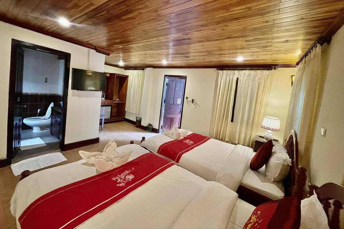 Habitación de hotel con dos camas, techo de madera, baño con inodoro y lavamanos, y un televisor.