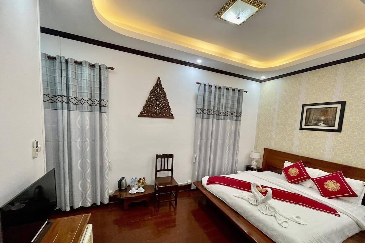 Una habitación de hotel con cama, una silla, una mesita con tetera y tazas, y un televisor.