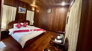 Habitación de hotel con techo de madera, cama con ropa de cama blanca y detalles rojos, mesita de noche con lámpara, una silla y una pequeña mesa con un control remoto y un menú.