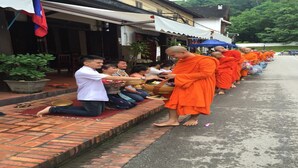 Monjes con túnicas anaranjadas realizando una limosna a un grupo de personas sentadas en una calle empedrada con ladrillos.