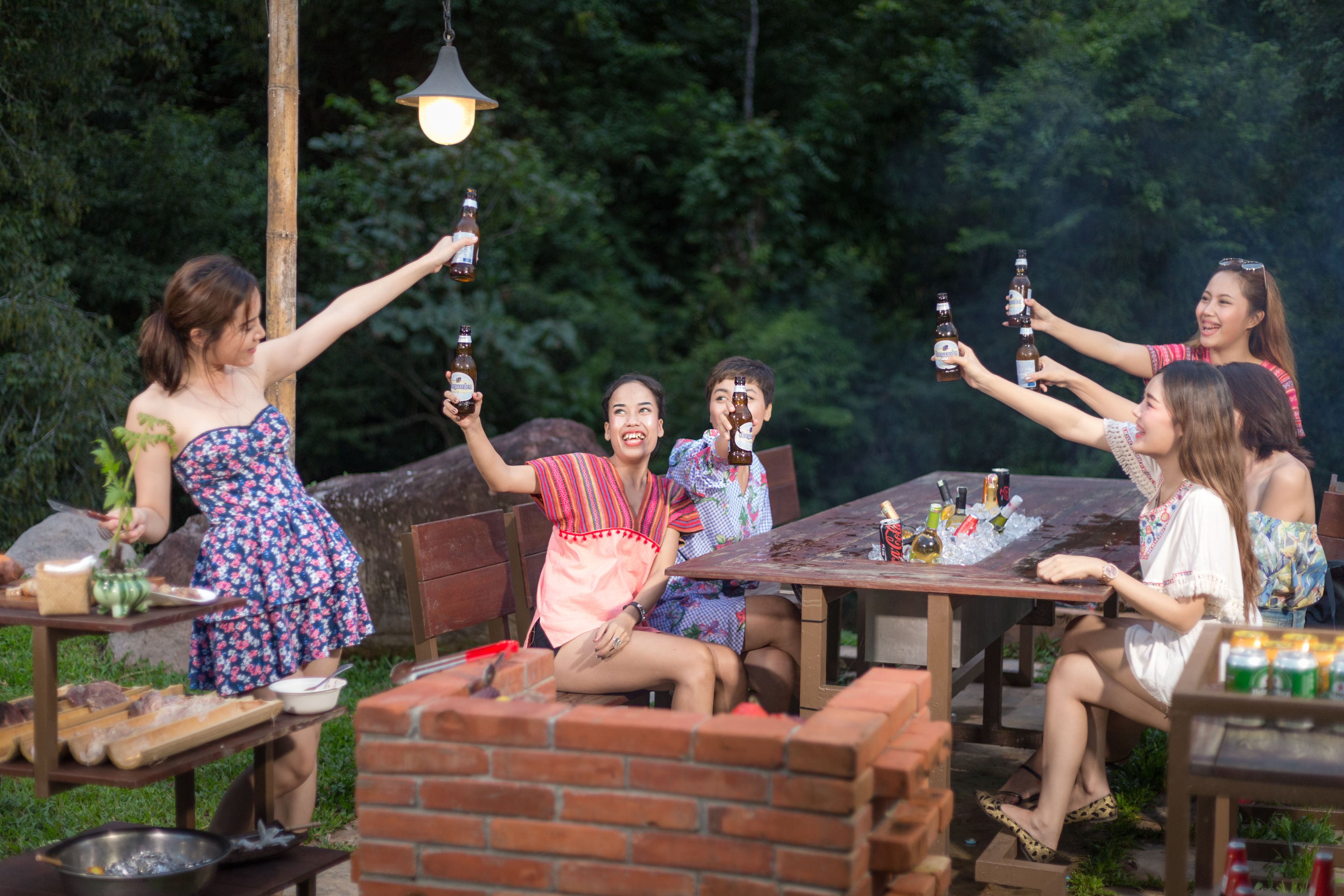 A group of people toasting with beer bottles at an outdoor gathering with a brick wall and greenery in the background.