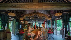 Un espace bar avec une table ronde centrale, entourée de chaises en osier avec des coussins rouges, et un plafond en bois agrémenté de luminaires suspendus.