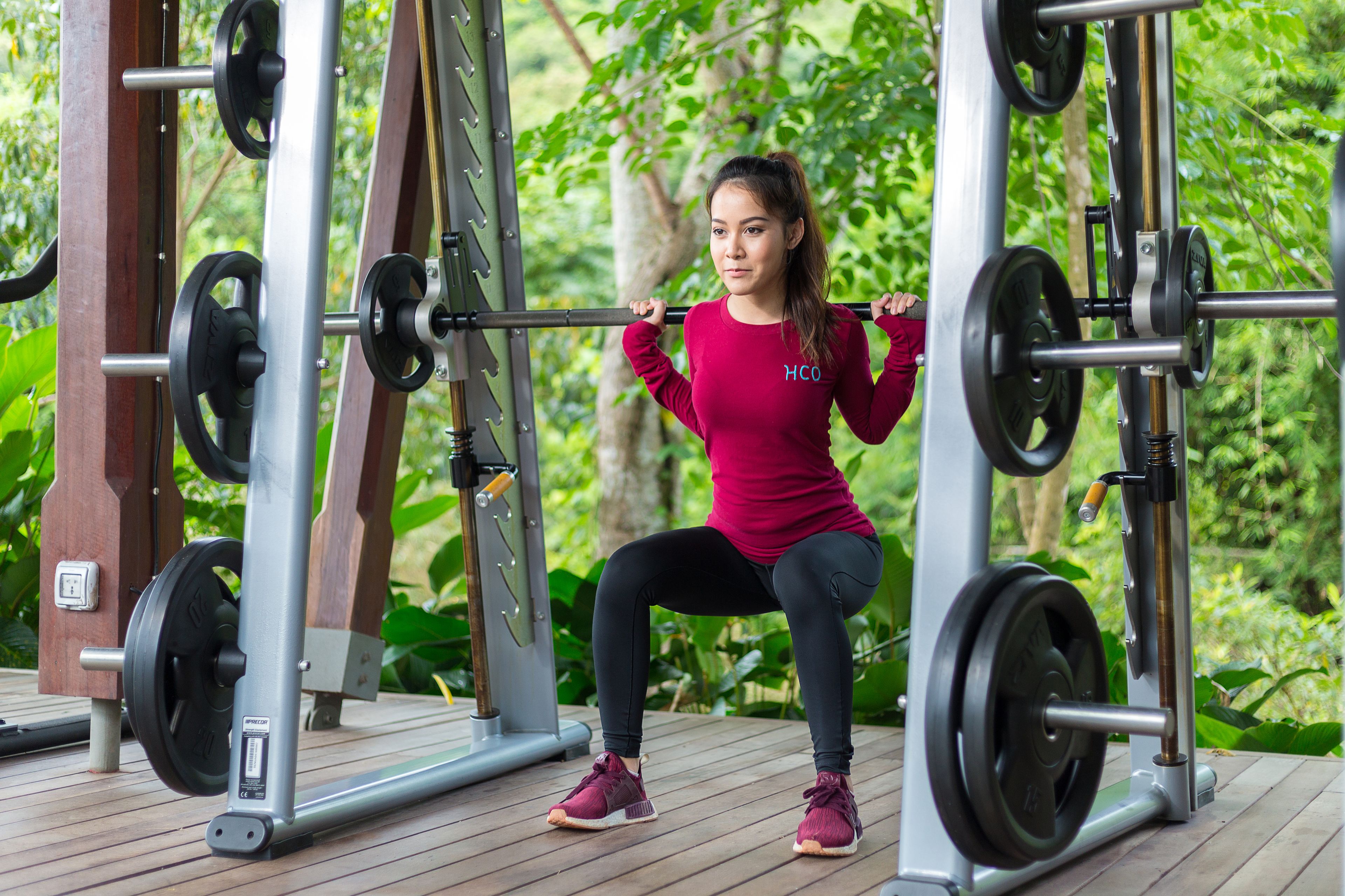 A woman performing squats on a gym machine outdoors with greenery in the background.
