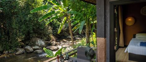 A room with a bed, a table with bottles, a camera, and a view of a stream and greenery.