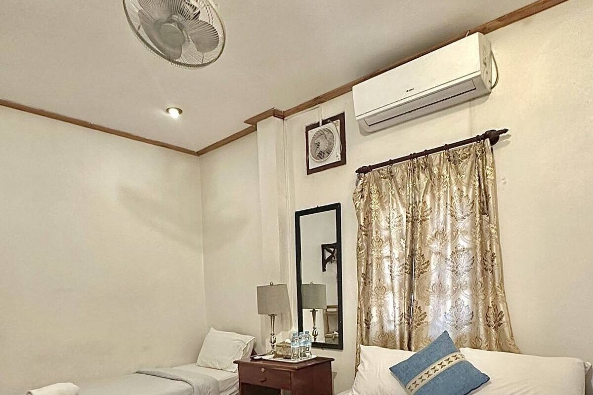 A hotel room with two single beds, a large bed, a nightstand with a lamp, an air conditioning unit, and a ceiling fan.