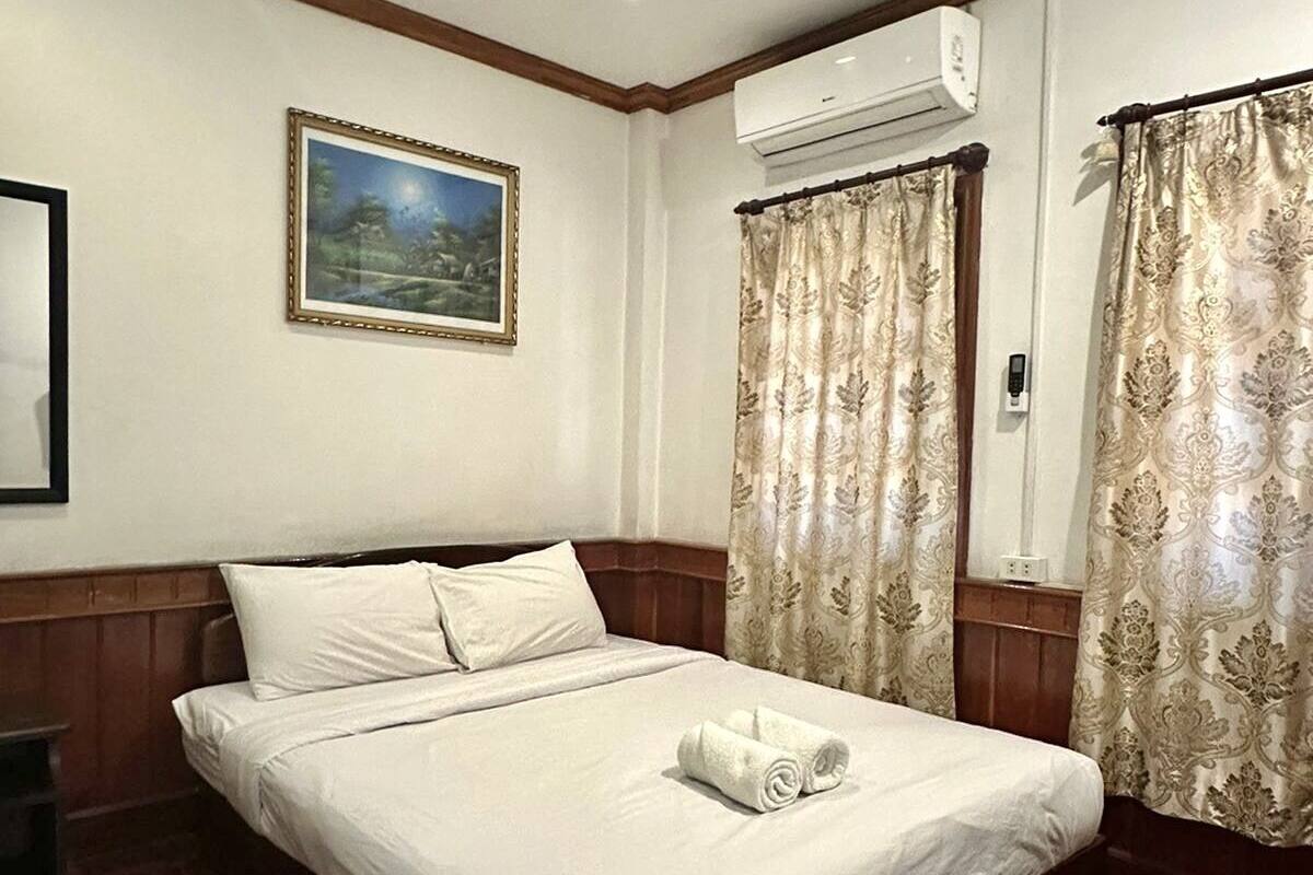 A neatly made bed with a rolled towel, a framed picture on the wall, and an air conditioning unit above it.