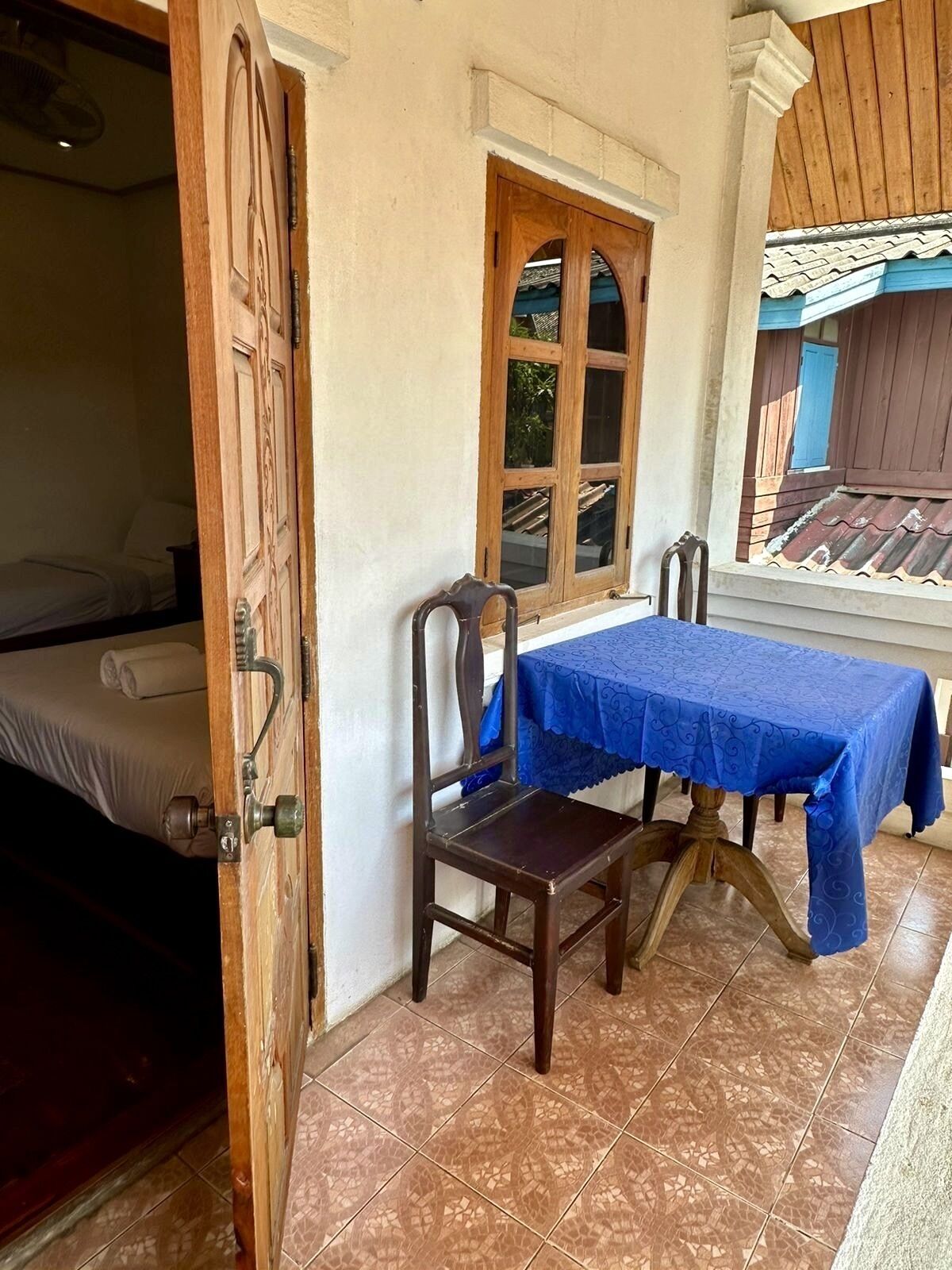 A room with a bed, a wooden chair, a table with a blue tablecloth, a window, and a tiled floor.