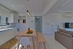 Una cocina moderna con armarios blancos, una mesa de comedor con sillas, un frutero y un sofá en una sala de estar con televisión.