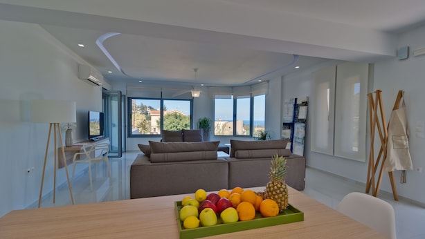 Una sala moderna con un gran ventanal, una mesa de comedor de madera con fruta y un sofá gris.