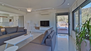 Una sala de estar moderna con un sofá, una mesa de centro y un televisor. Se aprecia un ventanal con vistas al exterior.
