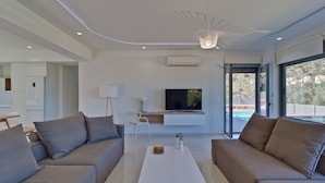 Un salón moderno con un sofá blanco, una mesa de centro, un televisor de pantalla plana y un amplio ventanal con vistas a una piscina y zonas verdes.