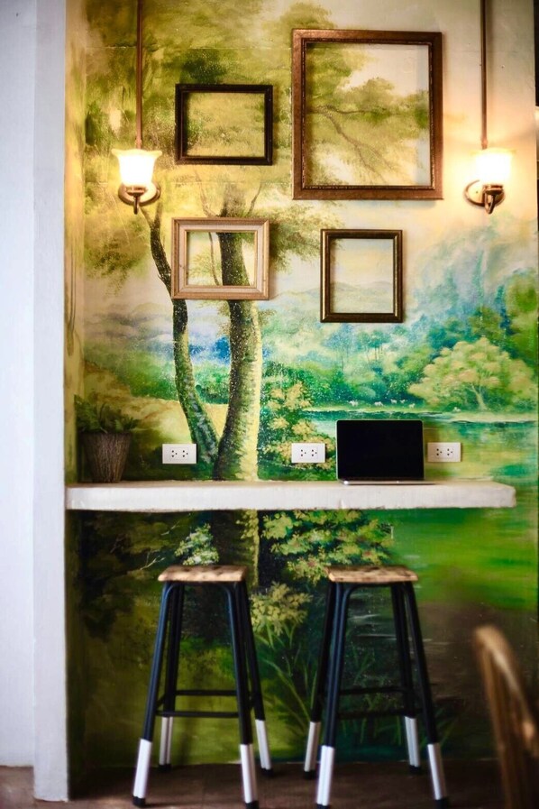 Una pared con un mural temático de naturaleza, dos cuadros enmarcados, una computadora portátil sobre una estantería y dos taburetes.