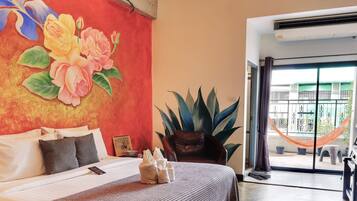 Une chambre d’hôtel avec un lit, une grande plante en pot, une vue sur le balcon extérieur et un mur rouge orné d’œuvres d’art florales.