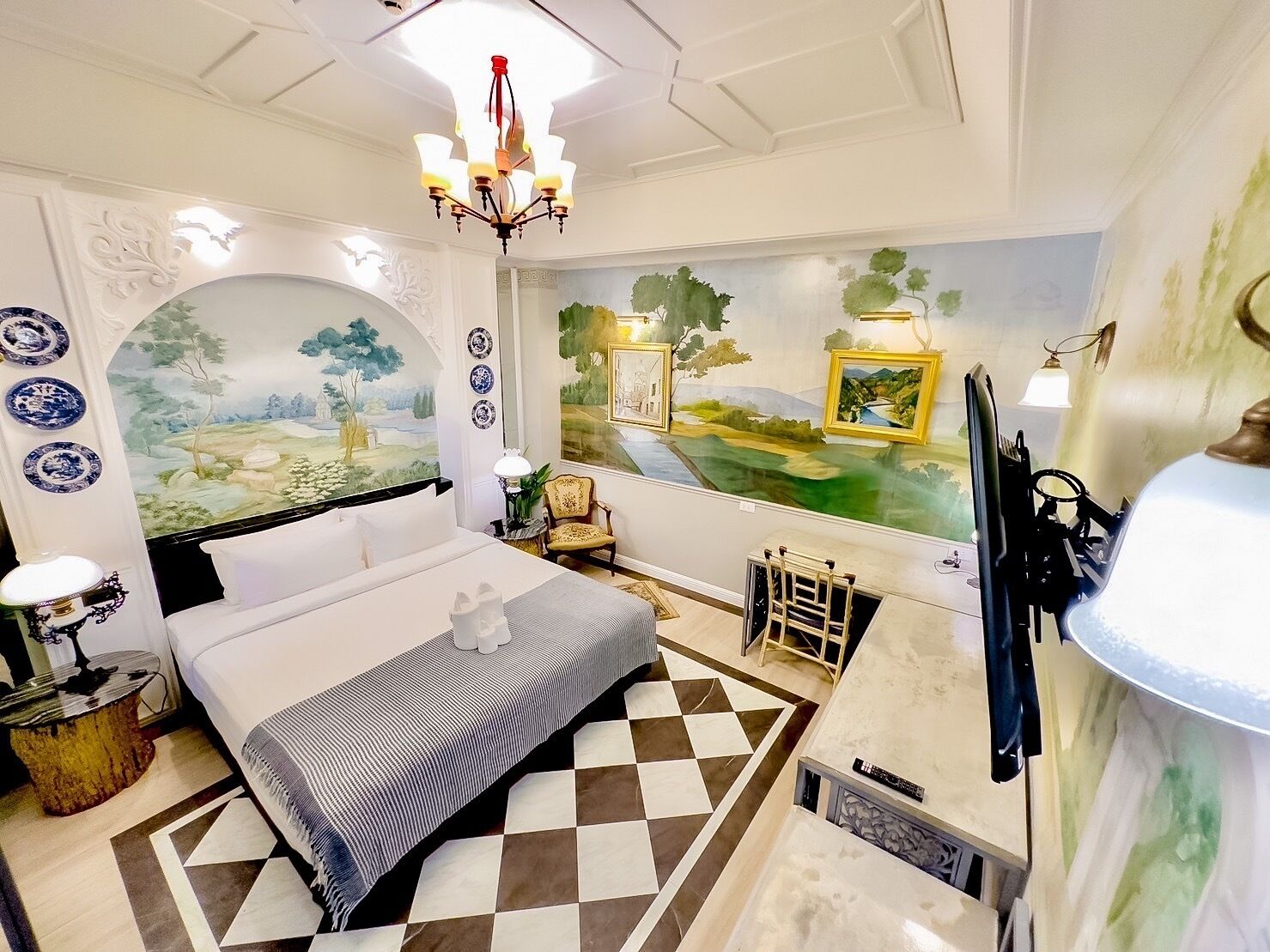 A hotel room with a bed, a desk, a chair, a chandelier, and a mural on the wall.