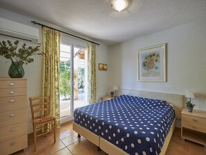 Een slaapkamer met een bed, een stoel, een dressoir, een nachtkastje, een lamp, een lijst met een afbeelding en een raam met gordijnen.