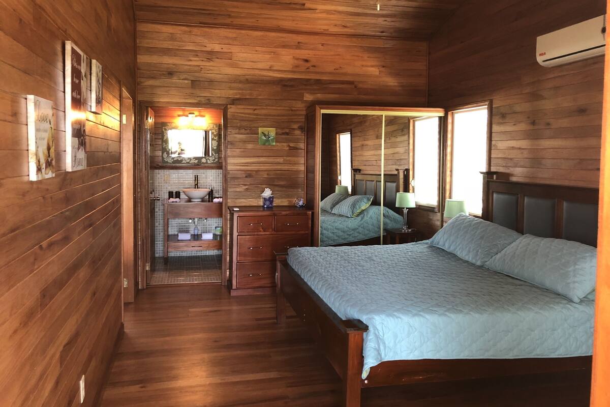 A wooden cabin bedroom with a bed, dresser, mirror, and a small bathroom area.